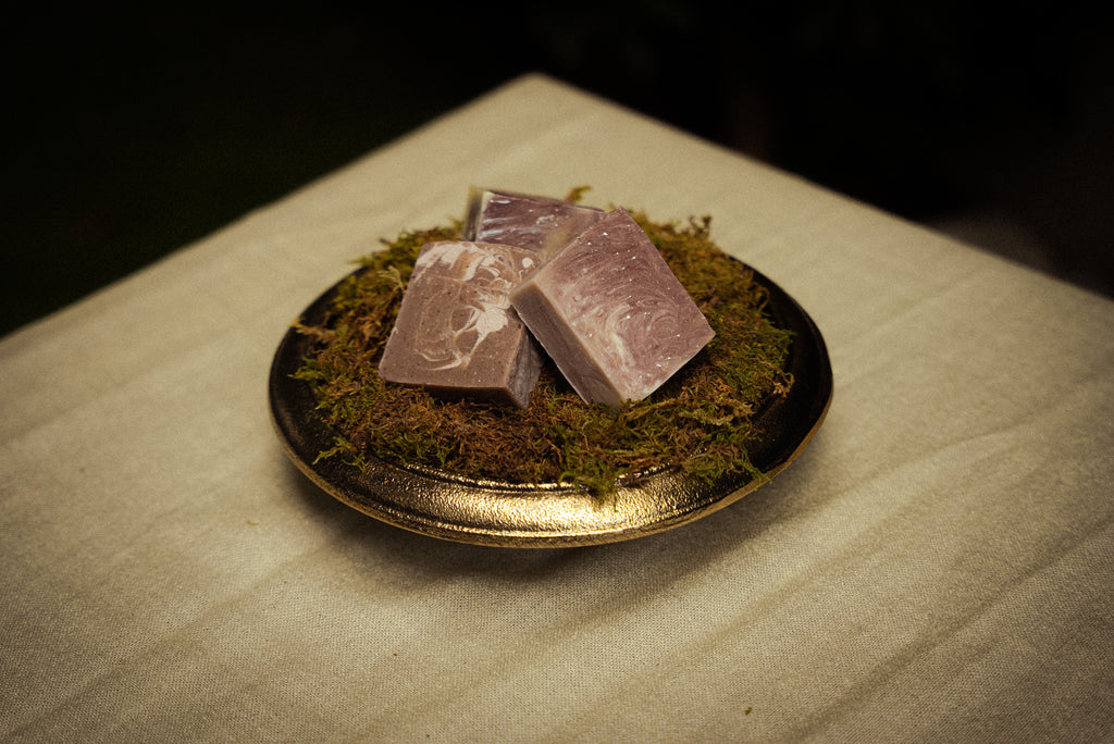 Three rectangular bars of lavender-colored soap with white swirls, resting on a bed of green moss inside a small gold-rimmed dish, set on a beige fabric surface.