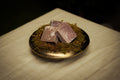 Three rectangular bars of lavender-colored soap with white swirls, resting on a bed of green moss inside a small gold-rimmed dish, set on a beige fabric surface.