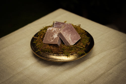 Three rectangular bars of lavender-colored soap with white swirls, resting on a bed of green moss inside a small gold-rimmed dish, set on a beige fabric surface.