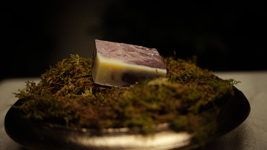A rectangular bar of handmade soap with a marbled purple and pale yellow design, resting on a bed of green moss inside a dark bowl.