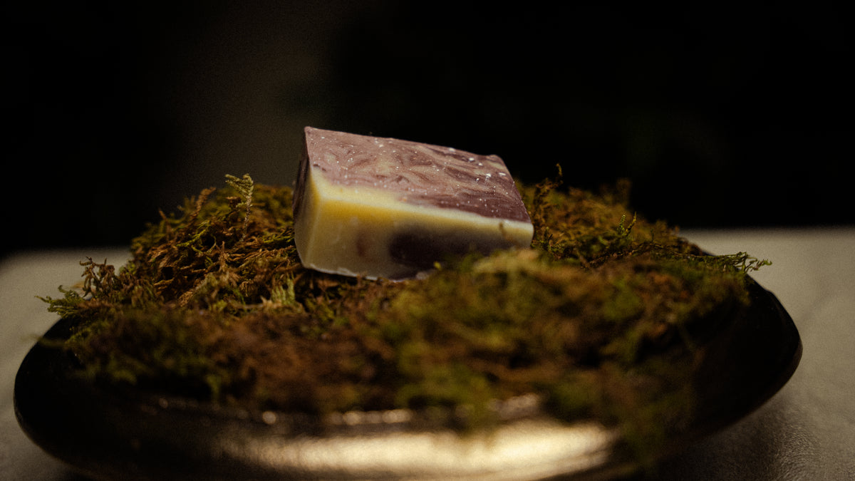 A rectangular bar of handmade soap with a marbled purple and pale yellow design, resting on a bed of green moss inside a dark bowl.
