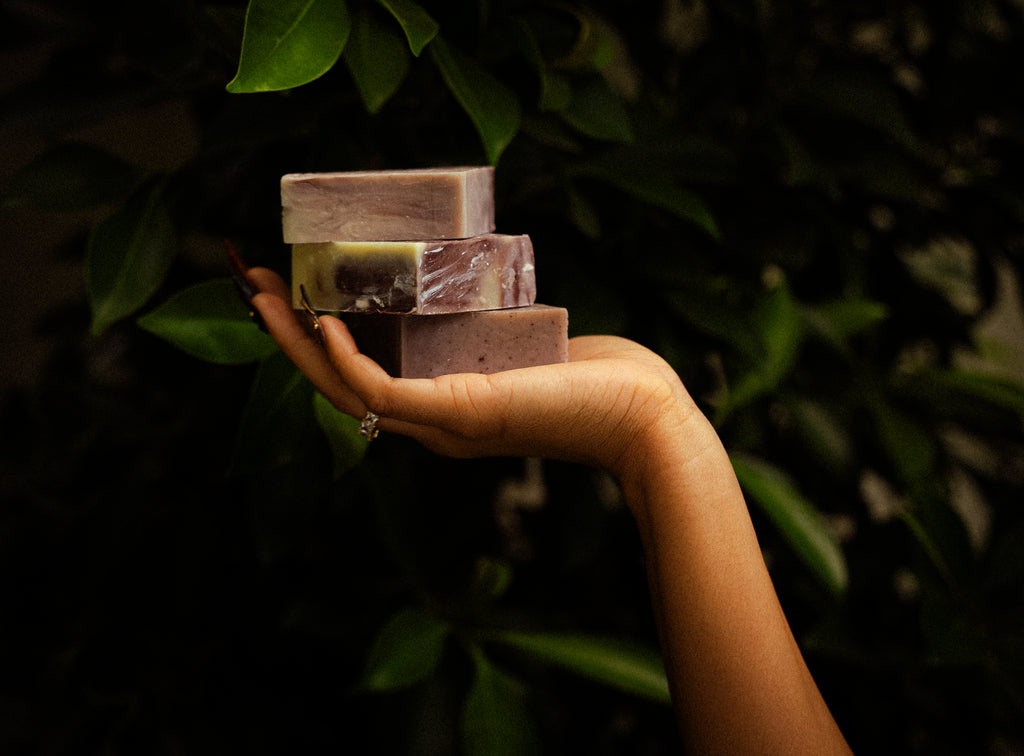 Three handmade rectangular bars of soap in earthy purple, olive green, and mauve tones stacked on a person's hand, set against a dark background of green leaves.