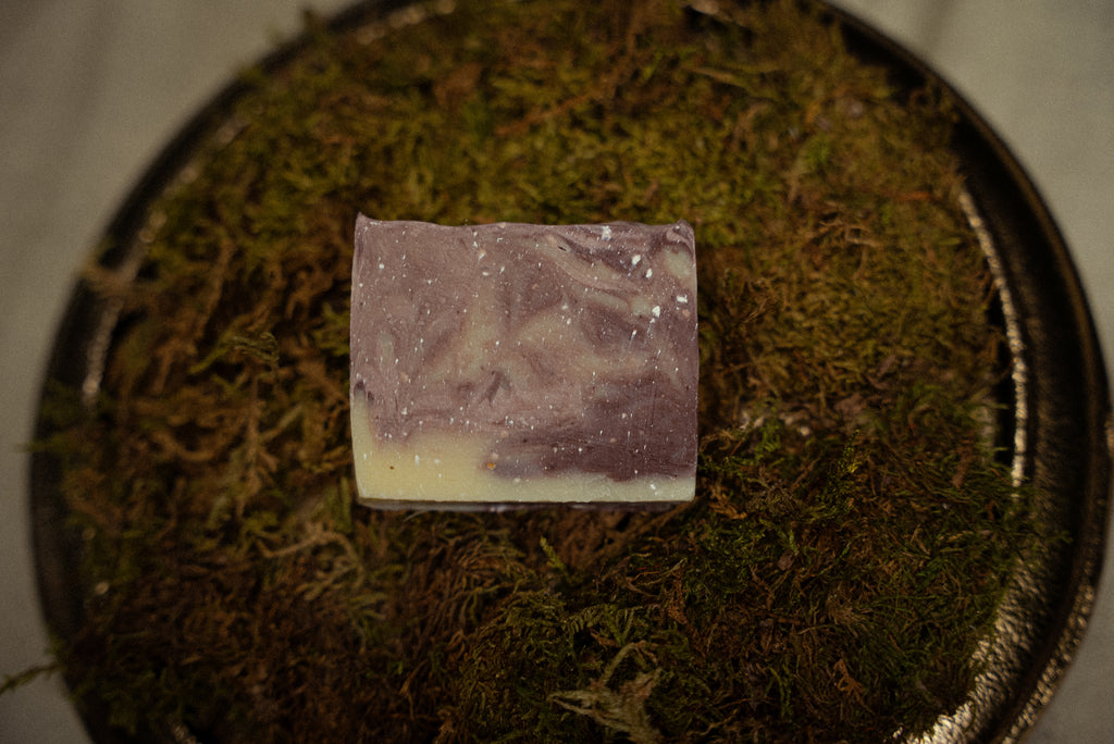 A rectangular bar of handmade soap with a lavender-purple and cream swirl pattern, resting on a bed of green moss inside a dark metal tray.