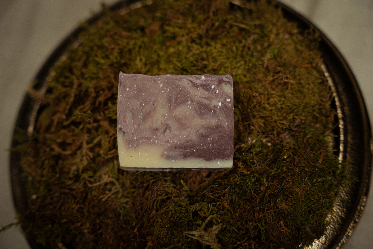 A rectangular bar of handmade soap with a lavender-purple and cream swirl pattern, resting on a bed of green moss inside a dark metal tray.