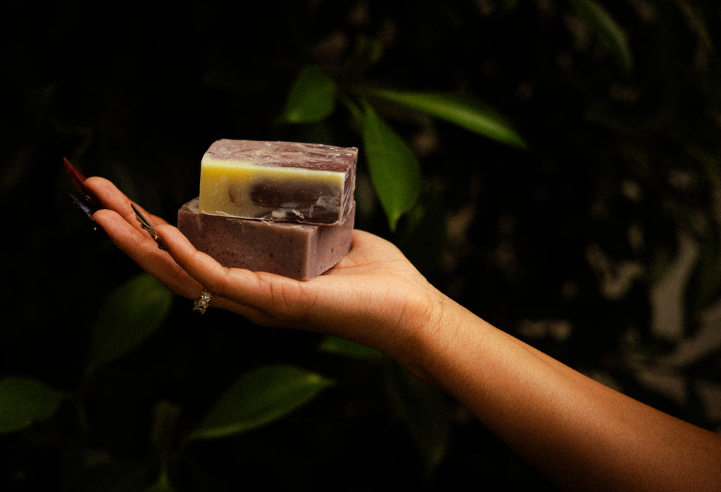 A hand holding two artisanal soap bars stacked vertically, with a dark purple outer bar and a yellow-and-white marbled inner bar, set against a dark, leafy background.
