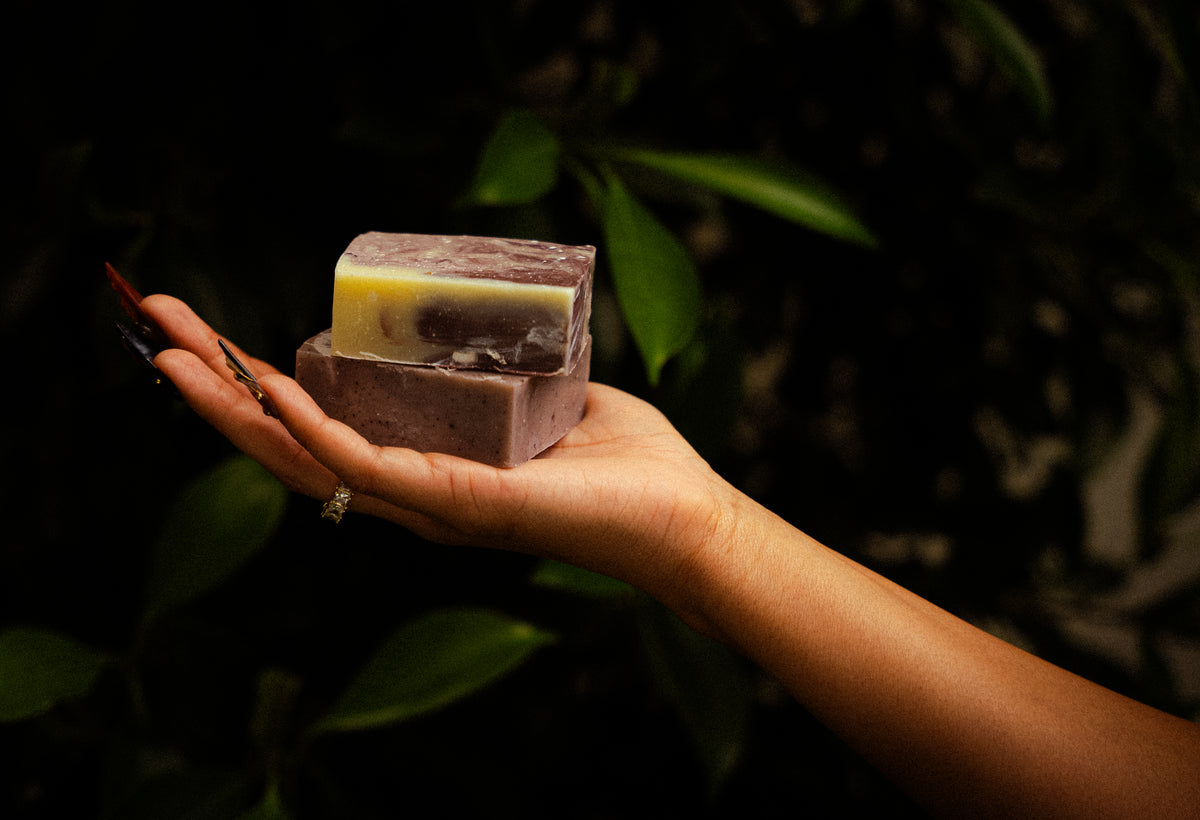 A hand holding two artisanal soap bars stacked vertically, with a dark purple outer bar and a yellow-and-white marbled inner bar, set against a dark, leafy background.