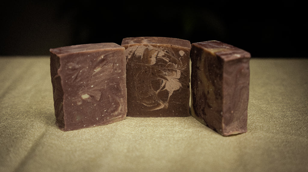 Three rectangular bars of handmade soap in a deep chocolate brown color, standing upright on a textured beige surface against a dark background.