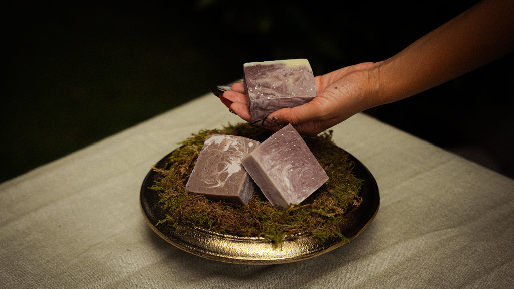 Three rectangular lavender-colored artisanal soap bars with white swirls are displayed on a gold-rimmed plate with green moss, with a hand holding one bar above the plate.